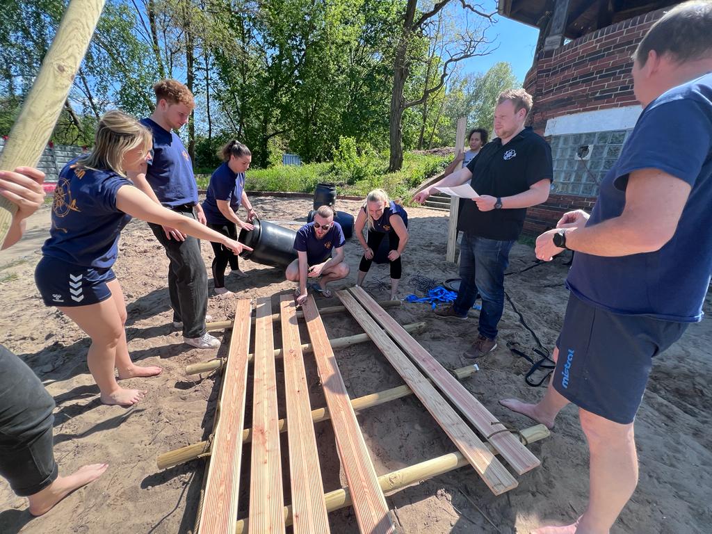 Eine Gruppe von vier Personen trägt gemeinsam ein Floß, das aus Holzlatten und Fässern gebaut ist, bei sonnigem Wetter in einer grünen Außenanlage.