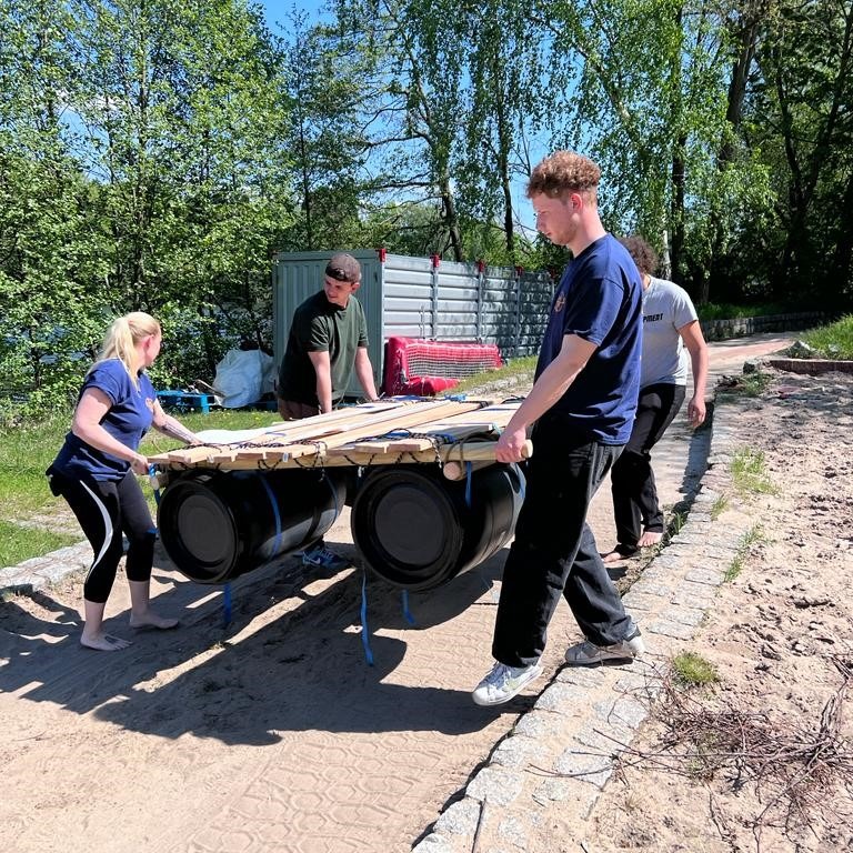 Eine Gruppe von vier Personen trägt gemeinsam ein Floß, das aus Holzlatten und Fässern gebaut ist, bei sonnigem Wetter in einer grünen Außenanlage.