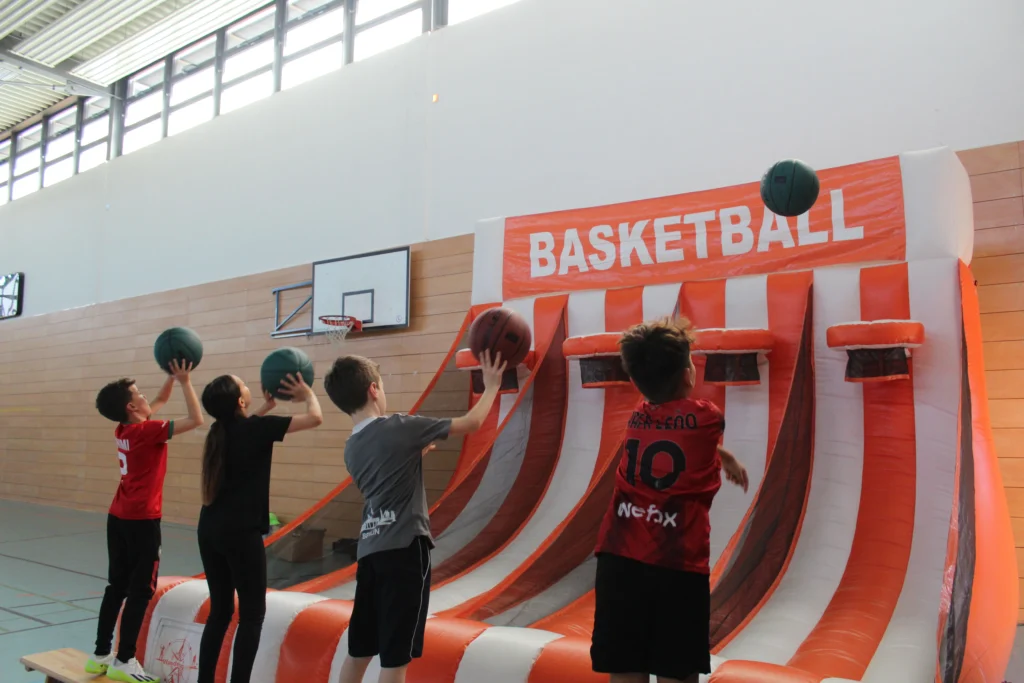 Mehrere Kinder werfen Basketbälle auf eine aufblasbare Basketballstation in einer Turnhalle, die mit einem großen orange-weißen Schriftzug 'BASKETBALL' gekennzeichnet ist.