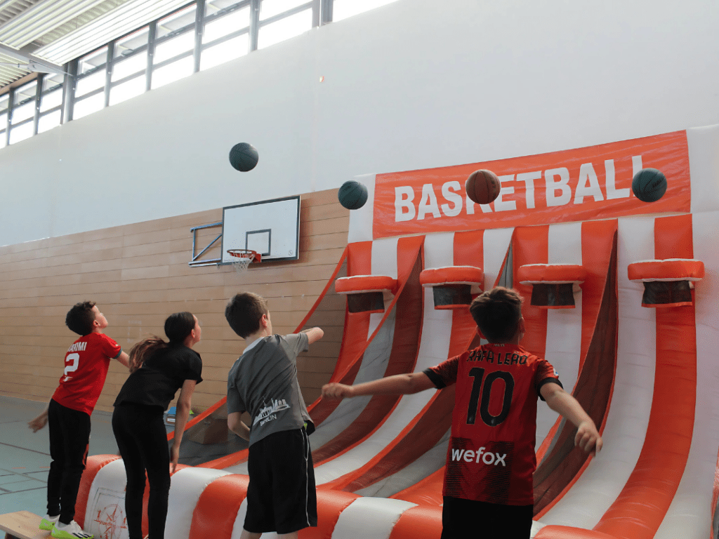 Mehrere Kinder werfen Basketbälle auf eine aufblasbare Basketballstation in einer Turnhalle, die mit einem großen orange-weißen Schriftzug 'BASKETBALL' gekennzeichnet ist.